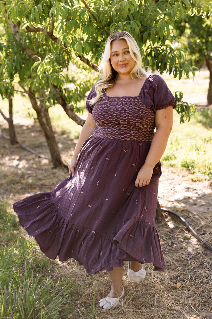 A woman with long blonde hair stands outdoors in a sunny, green area, smiling and holding the edge of her dress. She is wearing the Ivy City Co Acantha Dress in Aubergine Embroidered Gauze, which features puff sleeves and an elegant, maternity-friendly design.