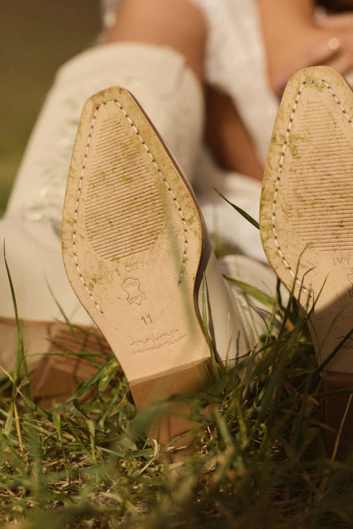 Close-up of the Avery Boot in Cream by Ivy City Co resting on grass, showing subtle grass stains and detailed stitching, with the wearer’s legs angled upward in the background.