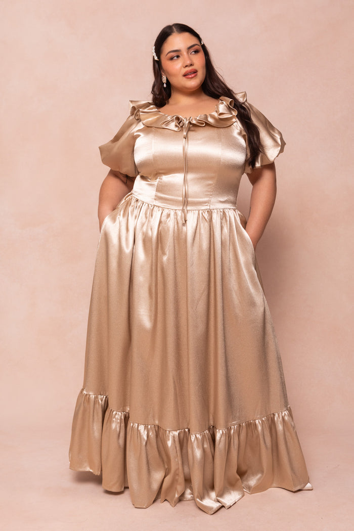 A woman in the Ivy City Co Billie Dress in Champagne Satin, with ruffled off-the-shoulder puff sleeves and a flared ruffled hem, stands with hands in her pockets against a beige background—ideal for bridesmaids.