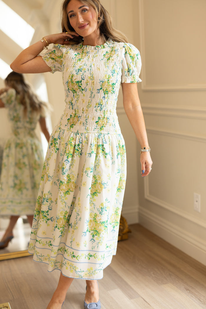 A woman smiles in a softly lit room, wearing the Ivy City Co Jennifer Dress in Garden Tea Party Floral. Her reflection appears in a nearby mirror, with light walls and wood floors setting a warm scene.