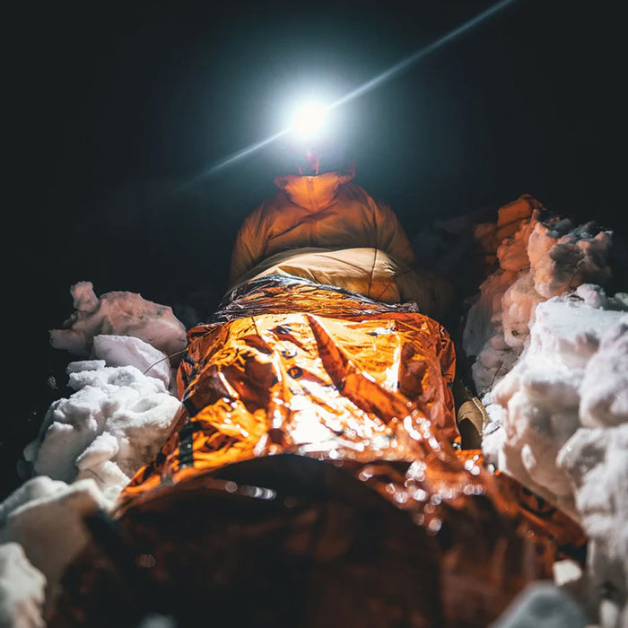 Lifestyle shot of a person with a head torch laying inside Lifesystems Heatshield Bivi Bag surrounded by snow