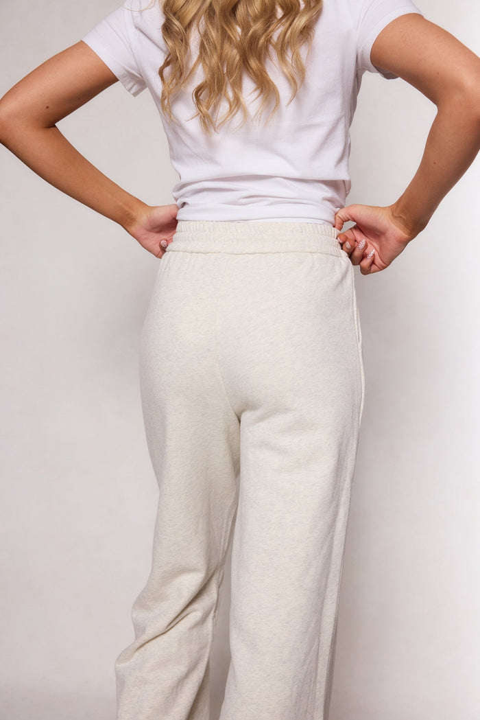 A person with long, wavy blonde hair stands with hands on hips, facing away from the camera against a light background, wearing a white short-sleeve shirt paired with the Ivy City Co Lisbeth Sweatpant in Oatmeal, featuring an elastic waistband.