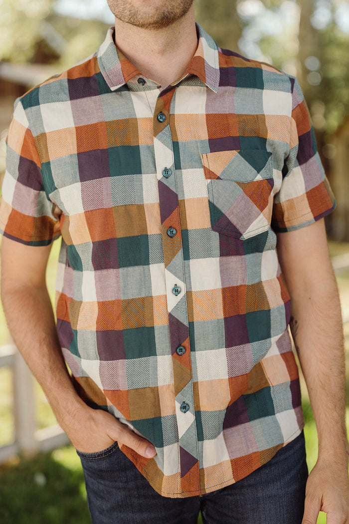 A person wearing the Ivy City Co Mens James Shirt in Maize Gingham, featuring short sleeves, a collar neckline, and a front pocket, stands outdoors with one hand in their pocket and blurred greenery in the background.