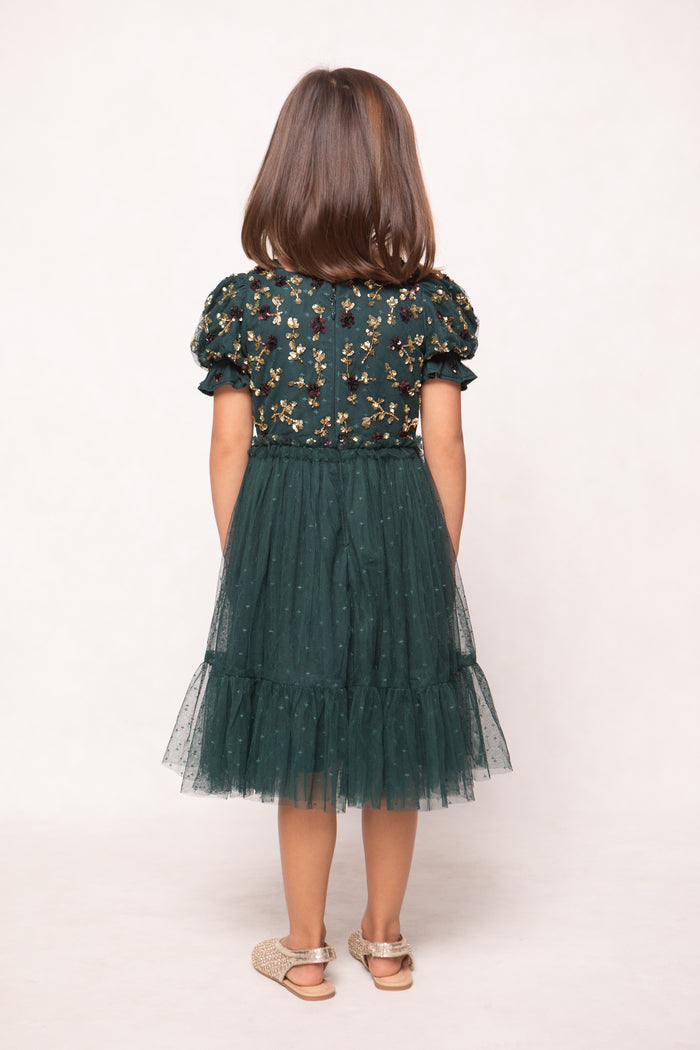 A young girl with straight brown hair faces away, wearing the Mini Adara Dress in Green by Ivy City Co, which features an embroidered bodice and tulle skirt, paired with gold sandals, set against a plain light background.