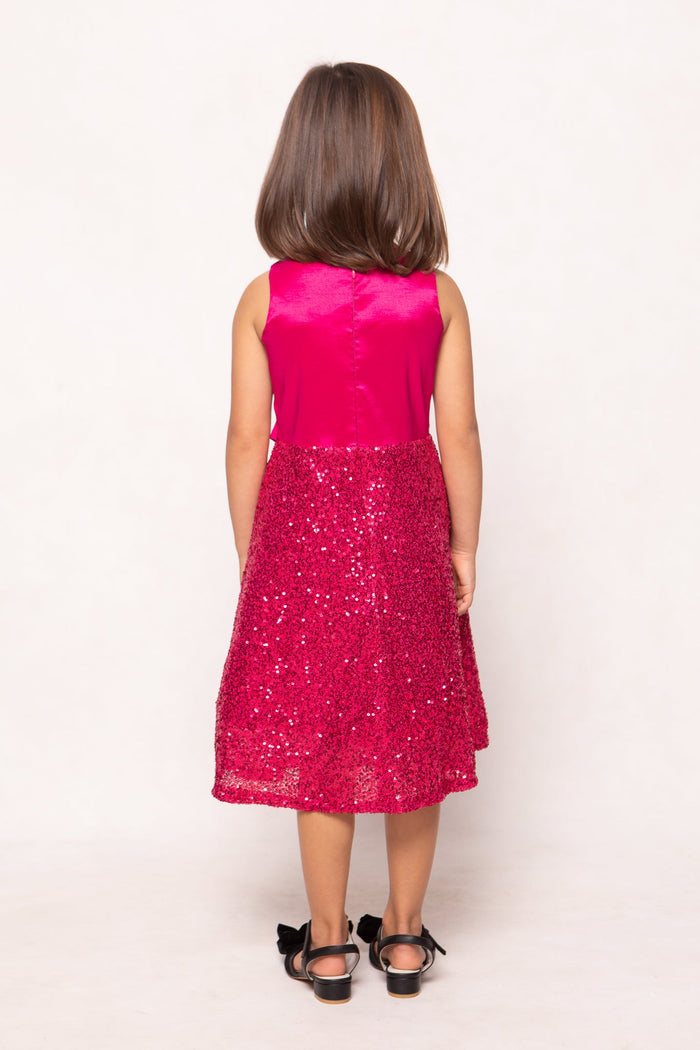 A young girl with straight brown hair stands facing away, wearing the sleeveless, knee-length Mini Alexis Dress in Fuchsia by Ivy City Co, featuring a shiny sequin skirt, paired with black sandals adorned with bows, against a plain light background.