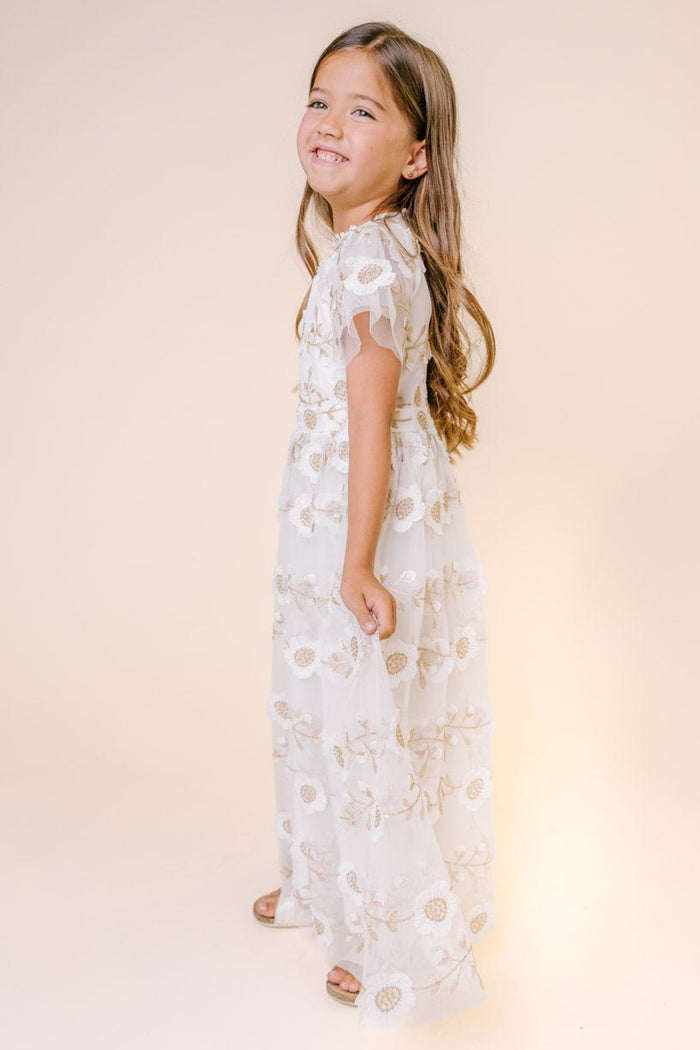 A young girl with long brown hair smiles while wearing the Ivy City Co Mini Athena Dress in White, an embroidered, floral-patterned maxi dress, as she stands sideways against a plain beige background.