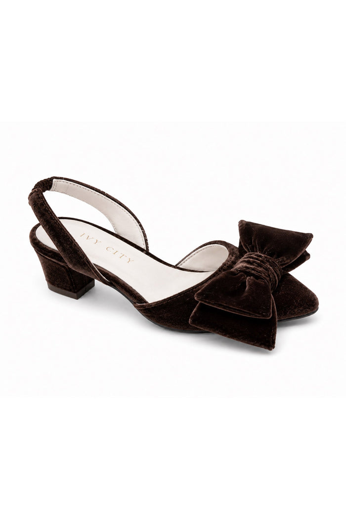 The Mini Eloise Bow Heel in Brown by Ivy City Co is a dark brown velvet slingback featuring a low chunky heel, a large bow detail on the toe, soft leather lining, and a sturdy rubber outsole, displayed on a white background.