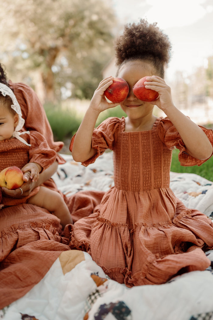 Two children in matching Ivy City Co Mini Madeline Dresses in Apple Cider sit on a picnic blanket; the older, wearing a dress with a charming smocked bodice and puff sleeves, playfully holds two apples over her eyes, while the younger holds an apple and looks down.