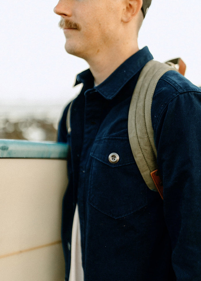Man in a blue jacket and backpack stands by a surfboard, gazing into the distance. Mustache and cap visible, ocean blurred behind.