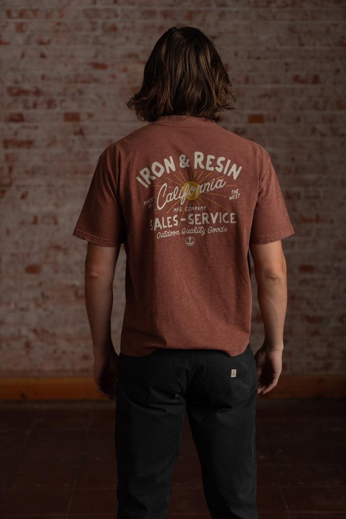 Man in brown "Iron & Resin California" shirt stands against a brick wall, showcasing back design. Casual, relaxed vibe.
