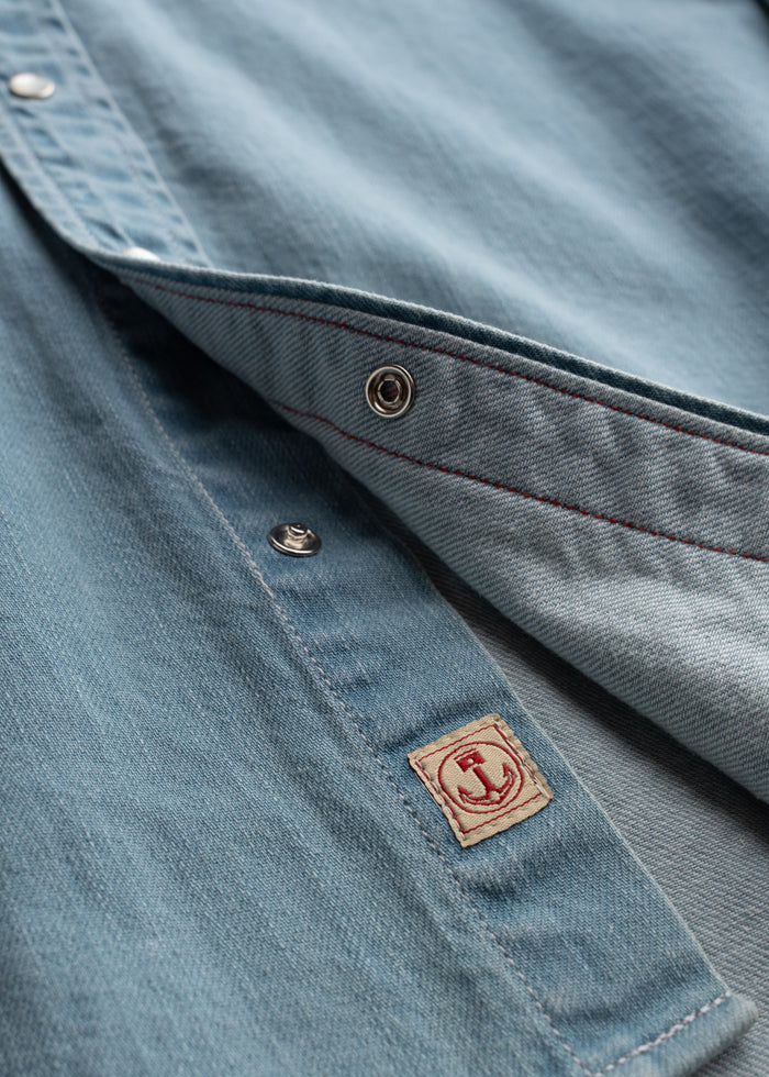 Close-up of a light blue denim shirt with metal snaps and a small anchor patch, highlighting texture and stitching details.