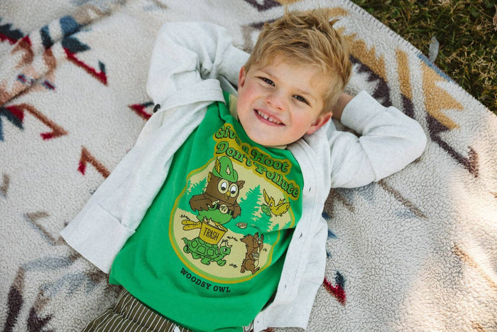 Boy relaxing on a blanket wearing a green Woodsy Owl and Friends youth ringer tee with a light gray hoodie.