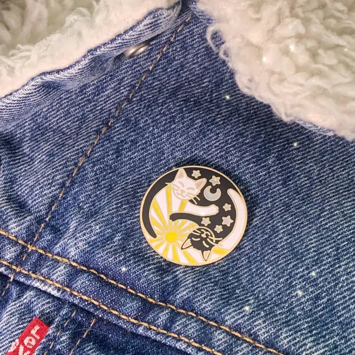 Round enamel pin featuring yin Yang cats with sun and moon motifs on blue denim.