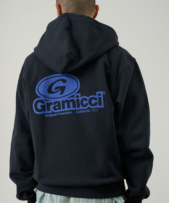 Back view of a model wearing a vintage black Gramicci '82 Zip Hooded Sweatshirt with a blue graphic and green pants on a light tone background.