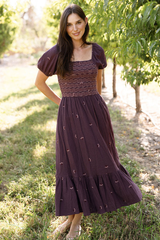 A woman stands barefoot on the grass in a sunlit orchard, smiling at the camera while wearing Ivy City Co’s Acantha Dress in Aubergine Embroidered Gauze—a maternity friendly, short-sleeve smocked midi dress with subtle puff sleeves and delicate embroidered details.