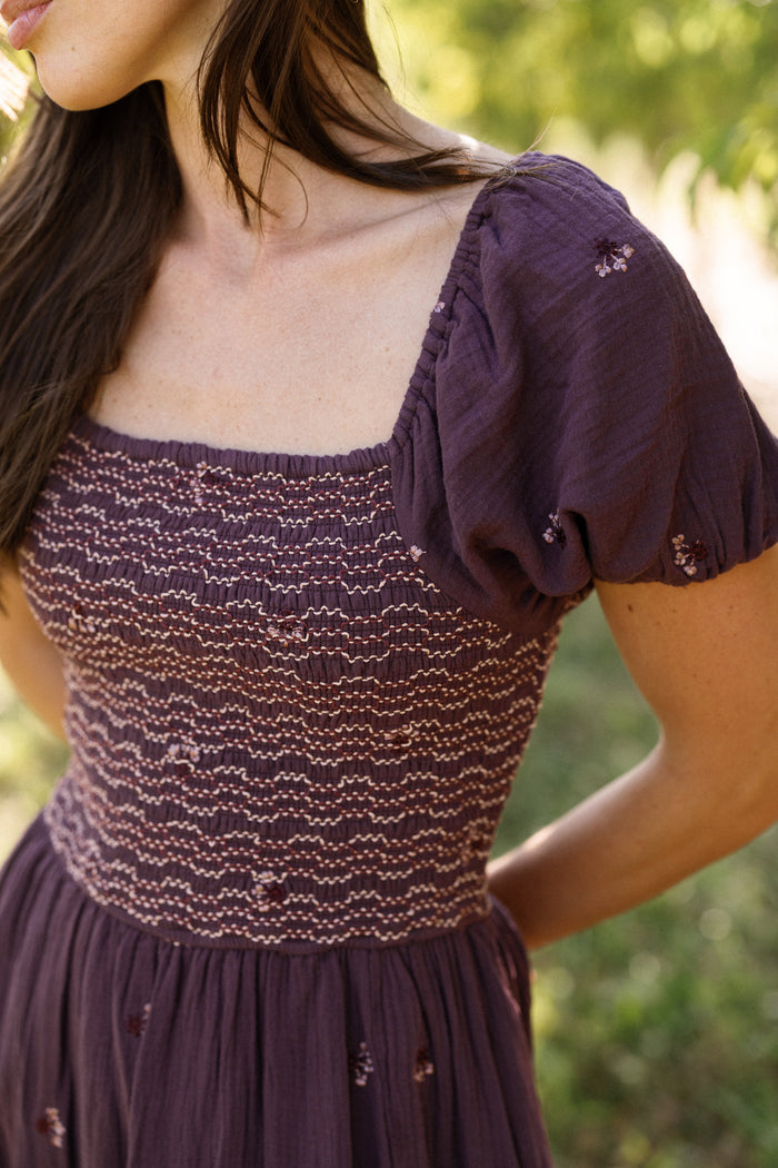 A woman wearing the Acantha Dress in Aubergine Embroidered Gauze by Ivy City Co—a dark purple smocked dress with puff sleeves, delicate floral embroidery, and a square neckline—poses outdoors amidst lush green foliage. This charming and maternity-friendly style is beautifully showcased in the natural setting.