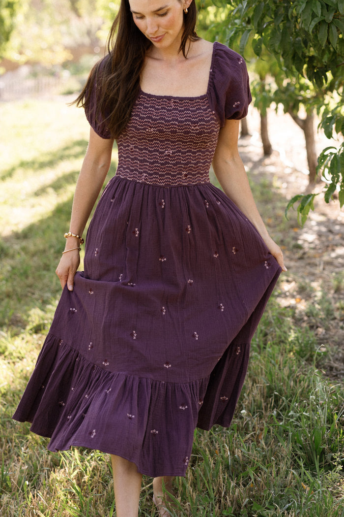 A woman stands outdoors on the grass, framed by trees and sunlight, wearing the Ivy City Co Acantha Dress in Aubergine Embroidered Gauze. The dress features elegant embroidery and charming puff sleeves. She holds out one side of her skirt and looks down, showcasing the beautifully maternity-friendly design that’s perfect for any stage.