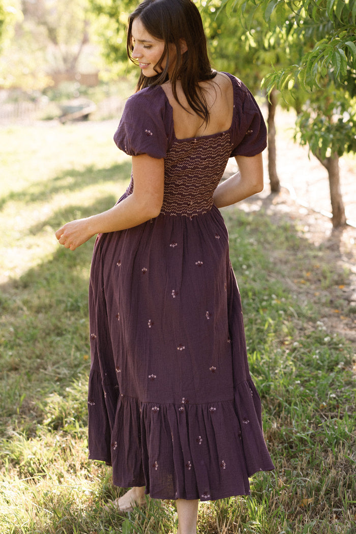 A woman in the Ivy City Co Acantha Dress in Aubergine Embroidered Gauze, featuring puff sleeves and a maternity-friendly fit, stands barefoot on grass, turning to the side with her back partially facing the camera, outdoors among sunlit trees.