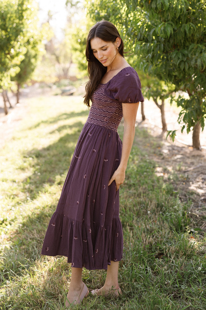Wearing the Ivy City Co Acantha Dress in Aubergine Embroidered Gauze—a purple, short-sleeved style with subtle puff sleeves and a smocked bodice—a woman stands on grass in a sunlit orchard, looking down as she gently touches her skirt. This elegant dress is also maternity friendly, making it an ideal choice for warm days outdoors.