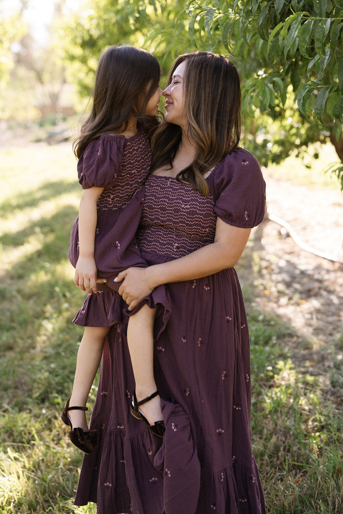 A woman holds a young girl as they smile at each other outdoors, both dressed in matching Ivy City Co Acantha Dresses in Aubergine Embroidered Gauze, featuring charming puff sleeves. Sunlight filters through green trees in the background, highlighting their maternity-friendly outfits.