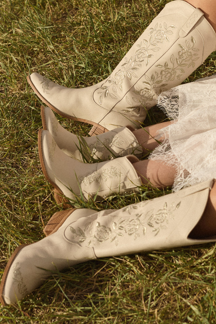 Two people sit on grass wearing the versatile Avery Boot in Cream by Ivy City Co. One, dressed in a white lace dress, shows only their legs and the embroidered boots for a stylish, coordinated look.