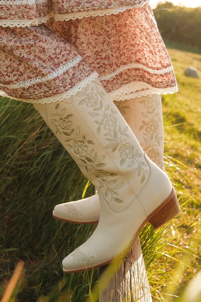 Wearing a floral pink and white dress, a person stands on a wooden fence in a sunlit grassy field, styled with the Avery Boot in Cream by Ivy City Co.