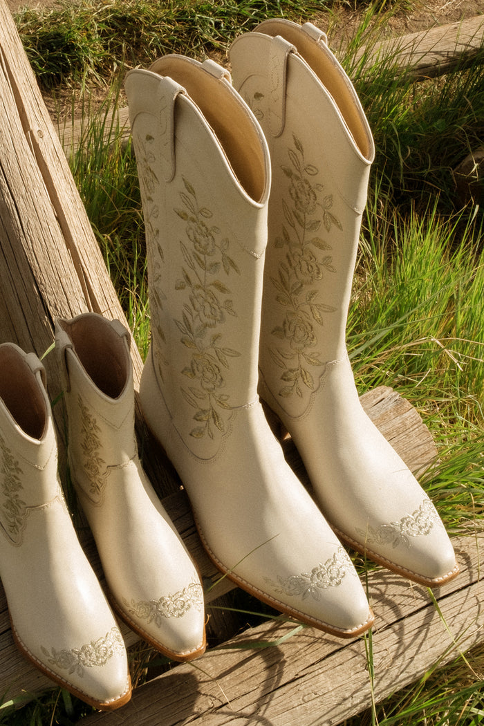 Three pairs of cream Ivy City Co Avery Boots with floral embroidery rest on weathered wooden planks beside tall green grass in sunlight.