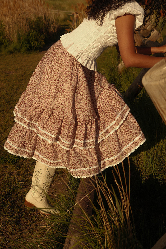 A woman in a white smocked top, long tiered floral skirt with lace trim, and the Ivy City Co Avery Boot in Cream leans on a wooden fence outdoors.