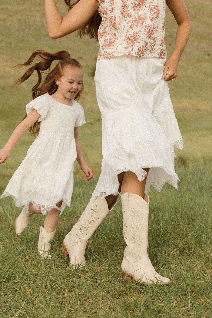A woman and a girl, visible from the waist down, joyfully run through a grassy field in white dresses and Ivy City Co's Avery Boot in Cream, with the girl's hair flowing as they move.