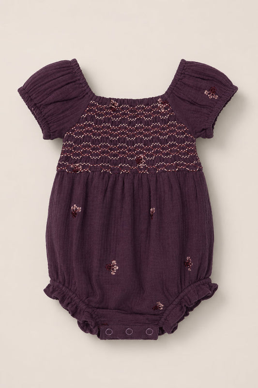 The Ivy City Co Baby Acantha Romper in Aubergine Embroidered Gauze features short puff sleeves, a smocked bodice adorned with delicate embroidered flowers, and convenient snap buttons at the bottom, shown laid flat on a light background.