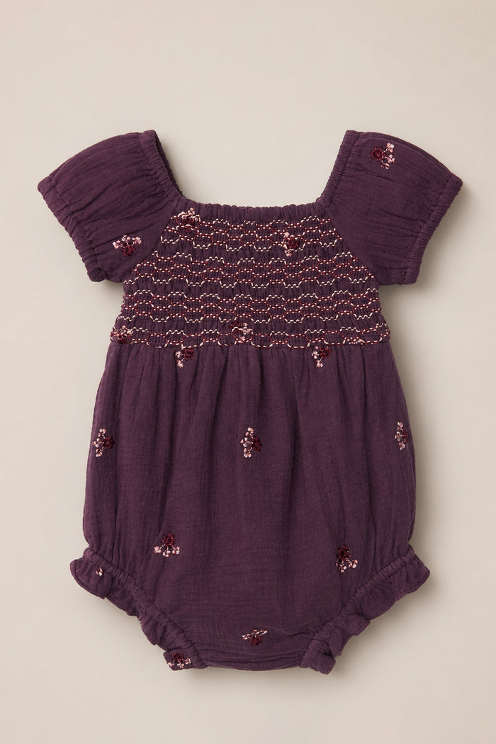 The Ivy City Co Baby Acantha Romper in Aubergine Embroidered Gauze showcases charming puff sleeves, a beautifully smocked bodice, elastic leg openings, and delicate floral embroidery scattered across the fabric. It is laid flat on a neutral background for a sweet and timeless presentation.