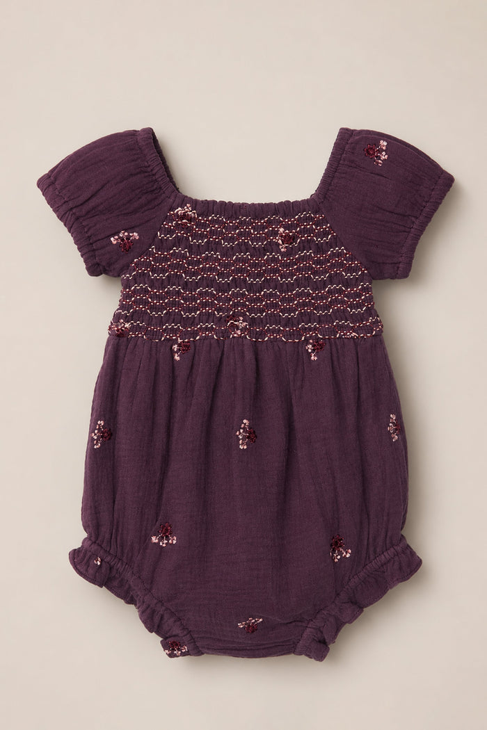 The Baby Acantha Romper in Aubergine Embroidered Gauze by Ivy City Co is a purple cotton romper featuring short puff sleeves, ruffled leg openings, a smocked chest, and delicate embroidered floral patterns scattered across the fabric.