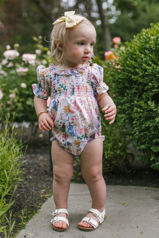 A young toddler with blonde hair, wearing the Ivy City Co Baby Cordelia Floral Romper, white sandals, and a yellow bow, stands on a garden path surrounded by green bushes and blooming flowers.