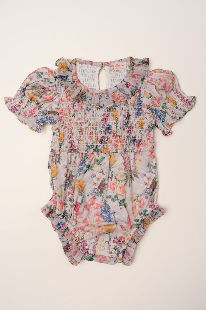 The Ivy City Co Baby Cordelia Floral Romper showcases a floral print with short puffed sleeves, ruffled trim, smocked bodice, and gathered elastic at the chest and legs, shown against a plain light background.