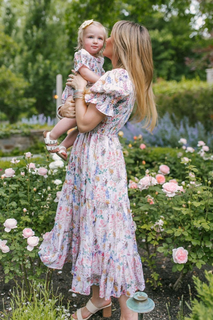A woman in a floral dress holds and kisses a young girl wearing the Ivy City Co Baby Cordelia Floral Romper, surrounded by pink roses and lush garden greenery.