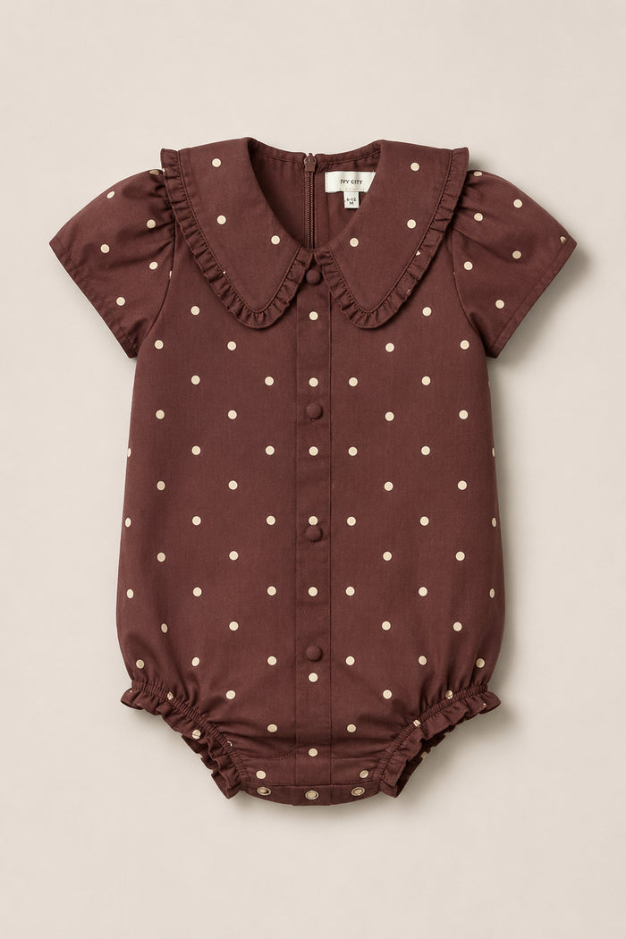 The Ivy City Co Baby Kit Romper in Warm Cocoa Dainty Dot features short puffed sleeves, a ruffled Peter Pan collar, white polka dots, and convenient snap buttons at the front and bottom. This charming cotton romper is displayed laid flat on a plain beige background.