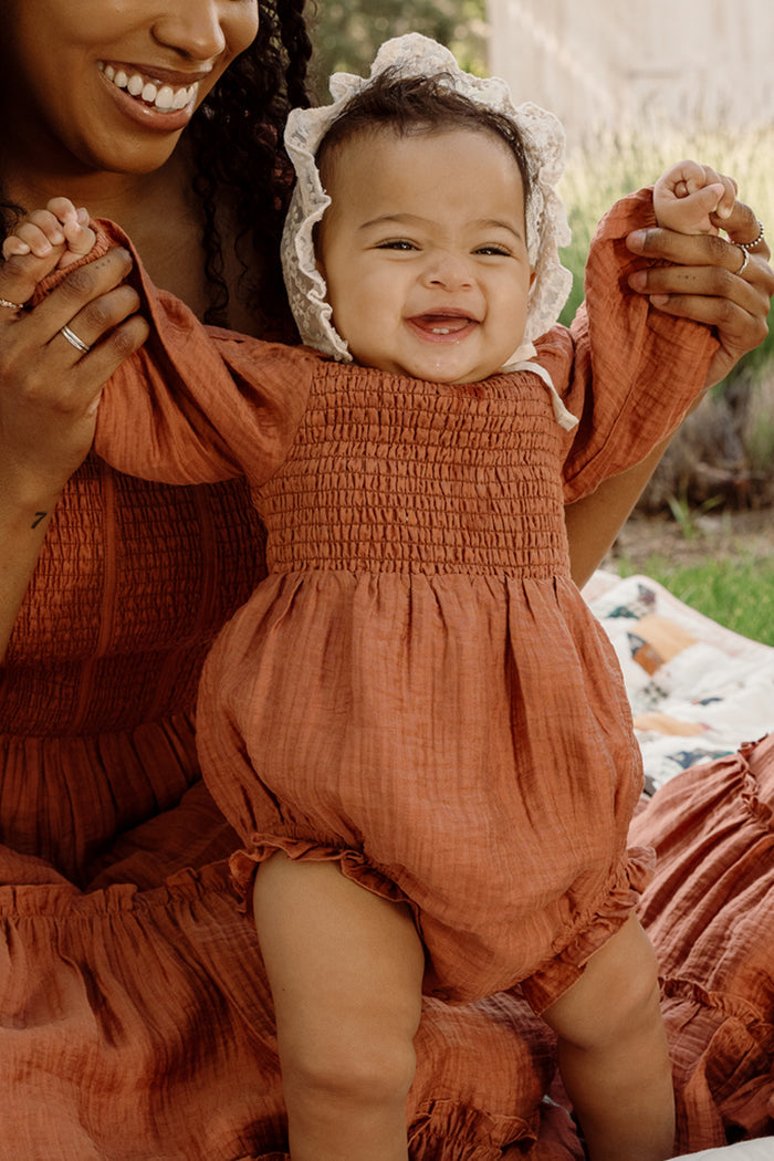 A smiling baby in the Ivy City Co Baby Madeline Romper in Apple Cider, featuring puff sleeves and a bonnet, is held up by an adult wearing a matching dress while outdoors on a blanket.