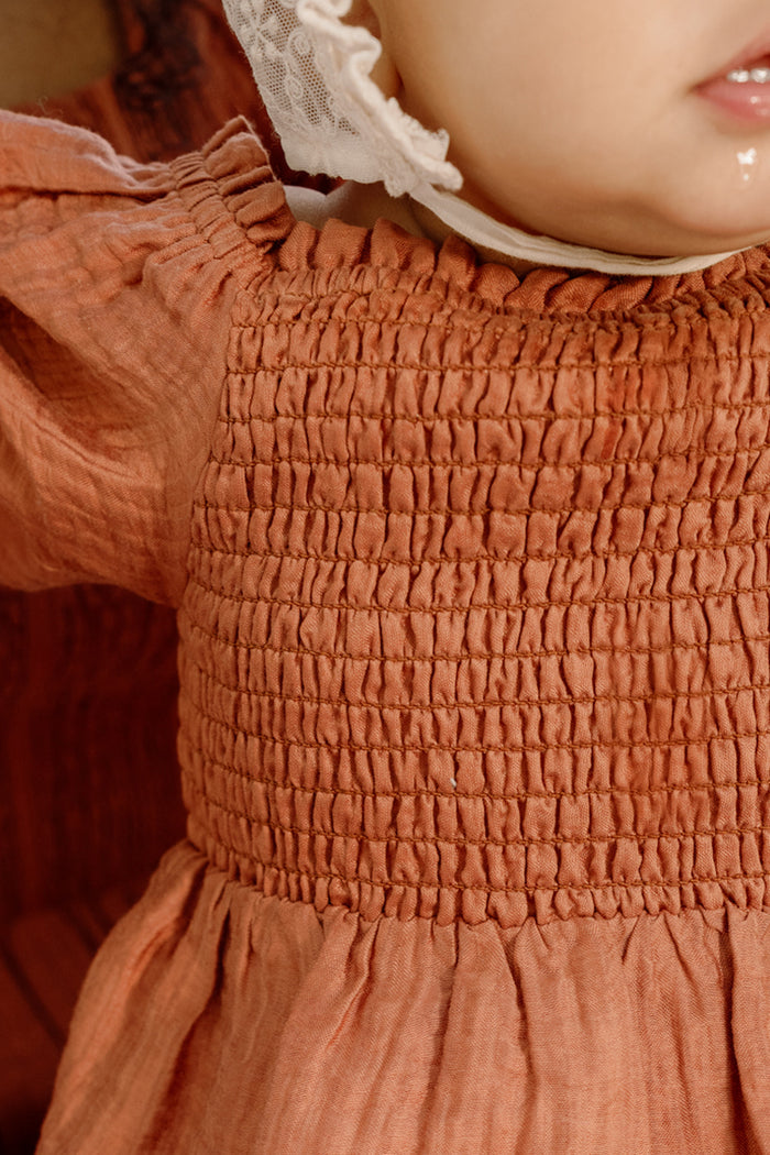 A close-up of a baby wearing the Ivy City Co Baby Madeline Romper in Apple Cider, featuring puff sleeves and a beautifully smocked bodice, paired with a white lace bonnet. Only the lower part of the baby’s face and upper body are visible.