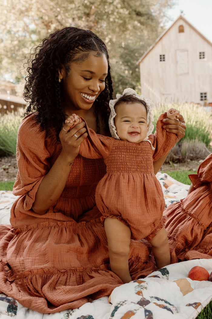 A woman in a rust-colored dress with puff sleeves smiles while holding a happy baby, who is wearing the adorable Ivy City Co Baby Madeline Romper in Apple Cider with a smocked bodice. They are sitting on a patterned blanket outdoors near a rustic building.