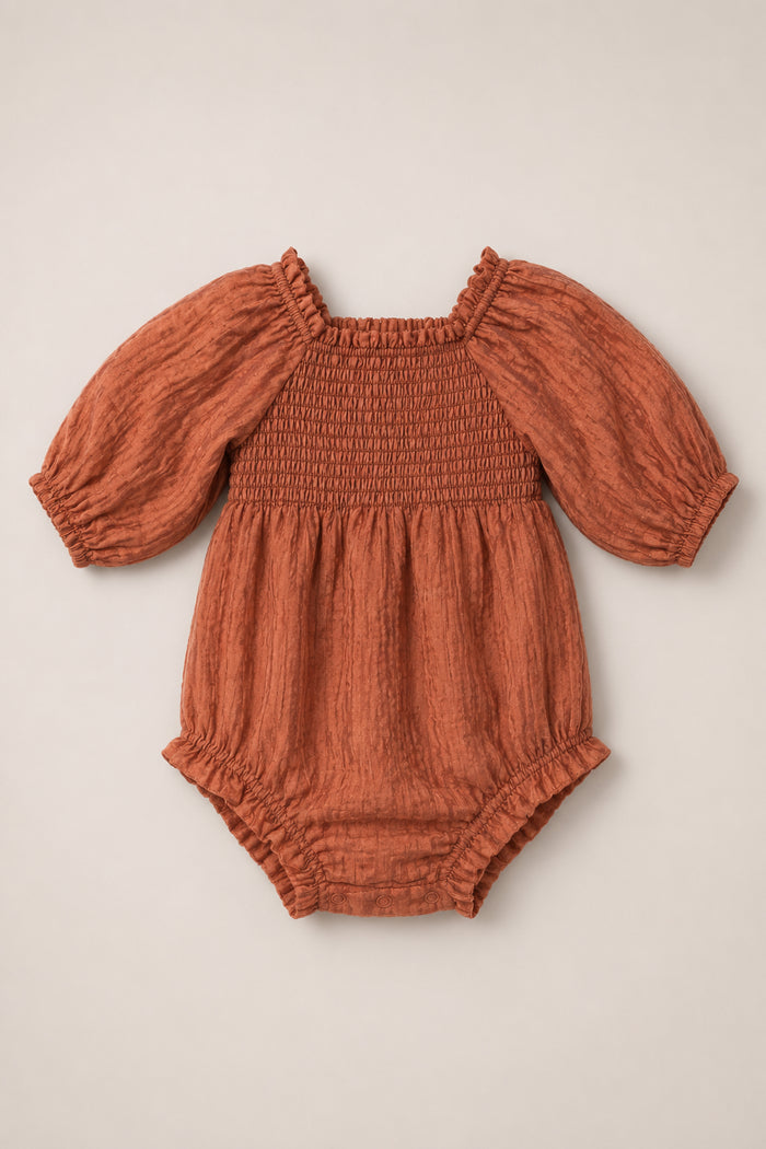 Ivy City Co’s Baby Madeline Romper in Apple Cider features a rust-colored long-sleeve design with a smocked bodice, subtle puff sleeves, elastic cuffs, and gathered leg openings, displayed on a neutral background.