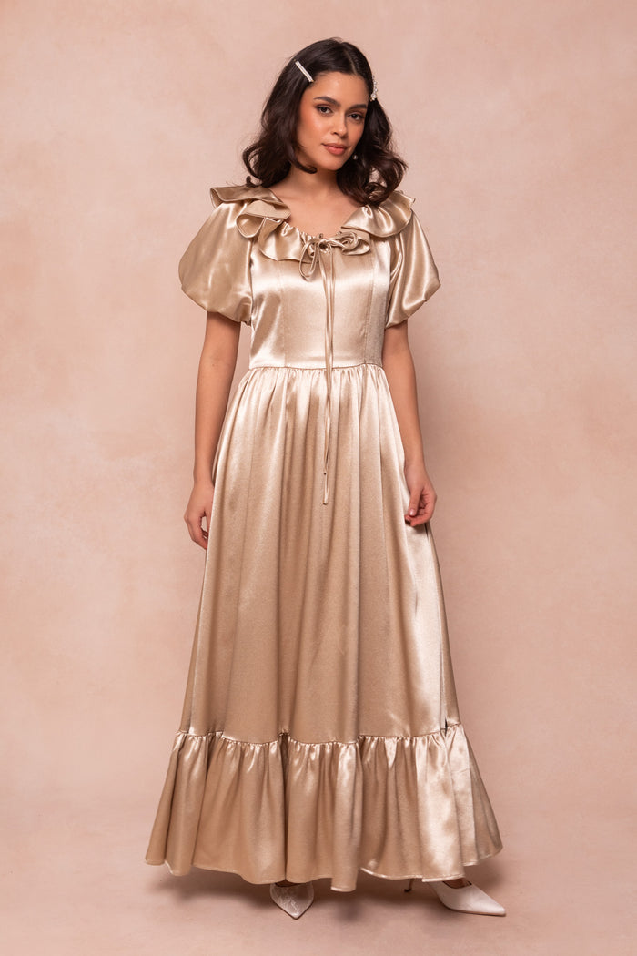 A woman with dark, shoulder-length hair and white pointed shoes stands against a light beige background, wearing the Ivy City Co Billie Dress in Champagne Satin—featuring short puff sleeves, a ruffled neckline, and a gathered waist.