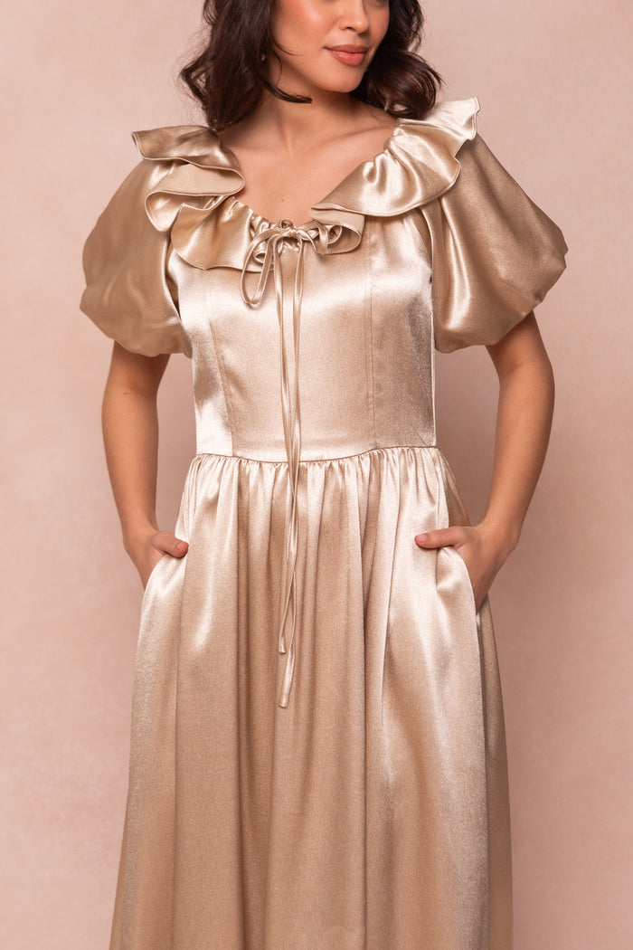 A woman wears the Billie Dress in Champagne Satin by Ivy City Co—a shiny, gold, short-sleeve dress with a ruffled neckline and puffed sleeves—standing with hands in pockets against a neutral background. Her face is partially out of frame.