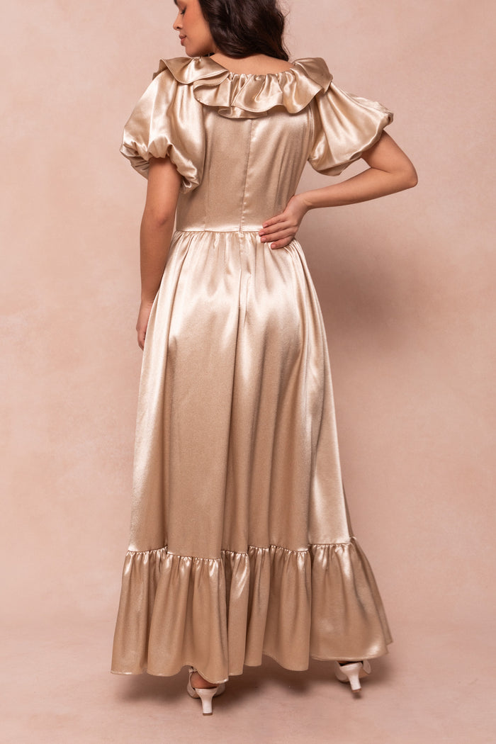 A woman in the Ivy City Co Billie Dress in Champagne Satin—a shiny, off-the-shoulder, puff-sleeve ruffled maxi with fitted bodice and flared skirt—stands with her back to the camera, hand on hip, against a beige background. Perfect for bridesmaids.