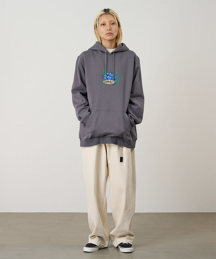 Model wearing the Gramicci Blueberry Hooded Sweatshirt with blueberry graphic in charcoal and cream colored pants on a light tone background.