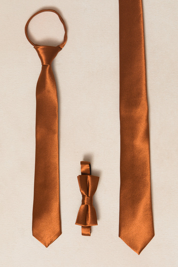 A flat lay of three rust-colored accessories on beige: a satin tie looped top left, a long necktie right, and Ivy City Co's Boys Max Tie in Amber Satin with adjustable strap at the bottom center.