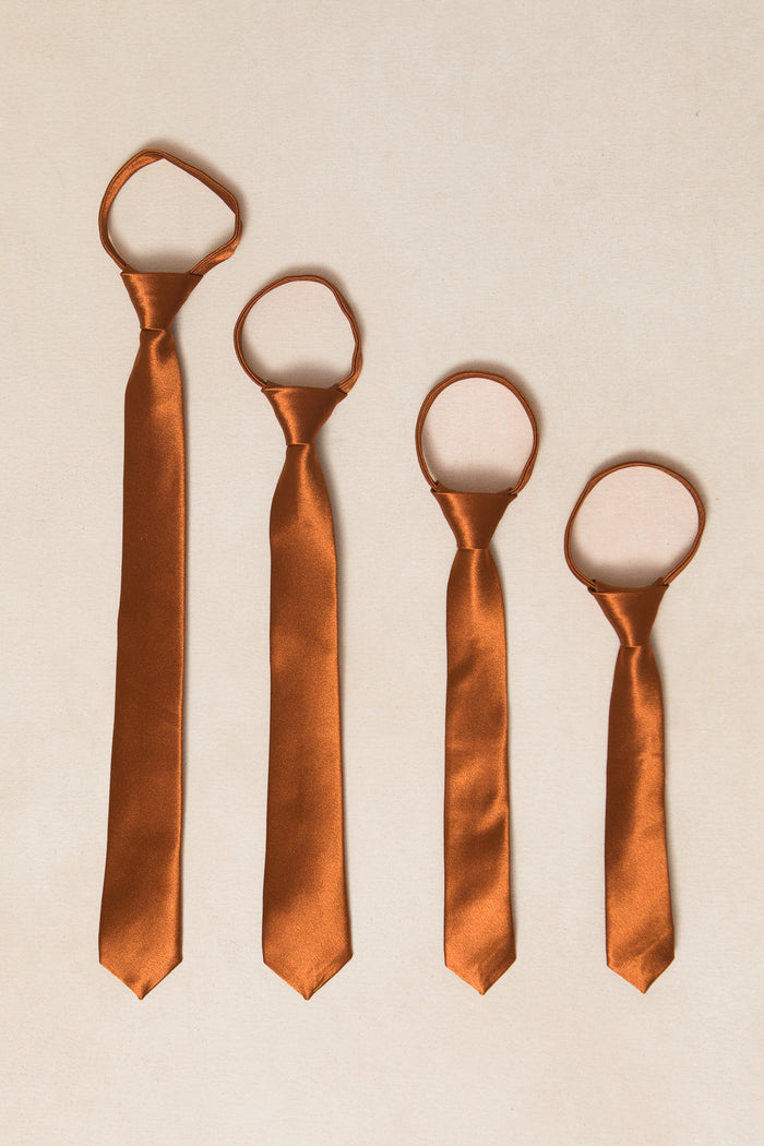 Four Ivy City Co Boys Max Ties in Amber Satin, arranged from shortest to longest on a light beige surface. Each tie is pre-tied with an adjustable zipper loop for easy wear.