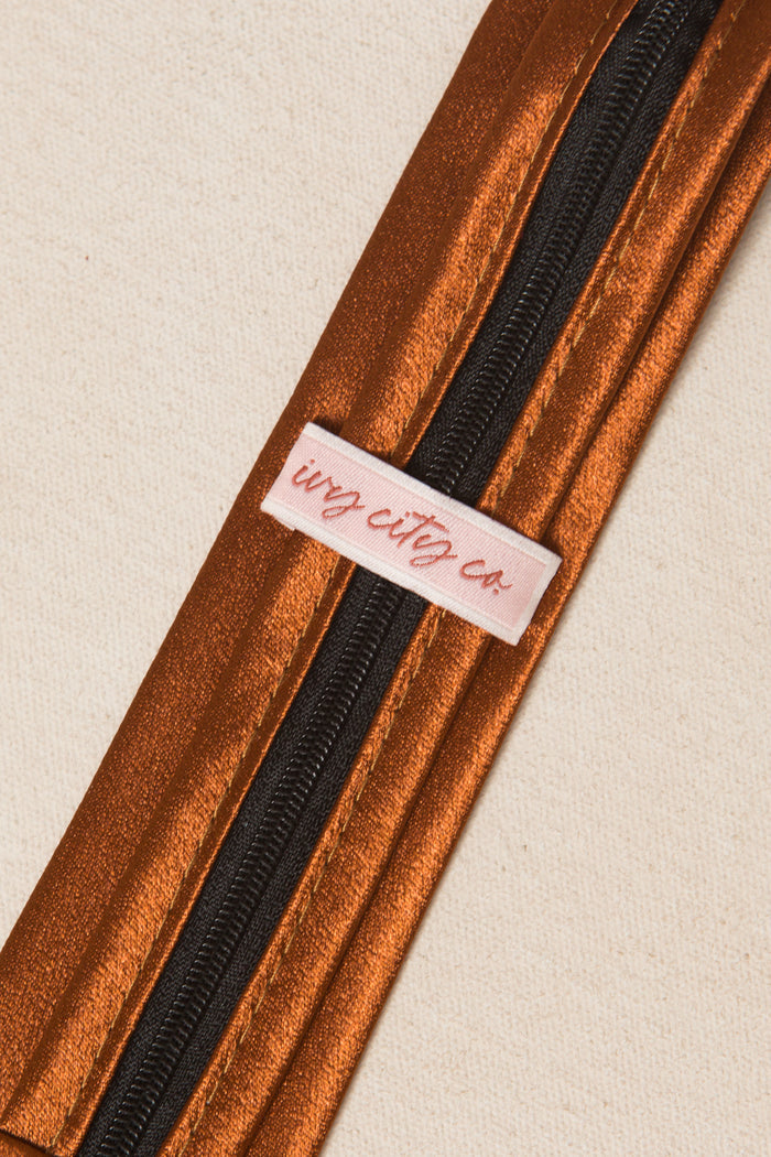 A close-up of the Boys Max Tie in Amber Satin by Ivy City Co, featuring a metallic copper adjustable zipper, black track, and a branded fabric label on a light beige background.