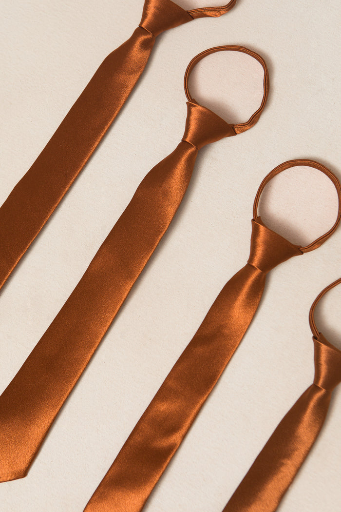 Four Boys Max Ties in Amber Satin by Ivy City Co, each featuring an adjustable zipper design, are arranged diagonally on a light beige surface.