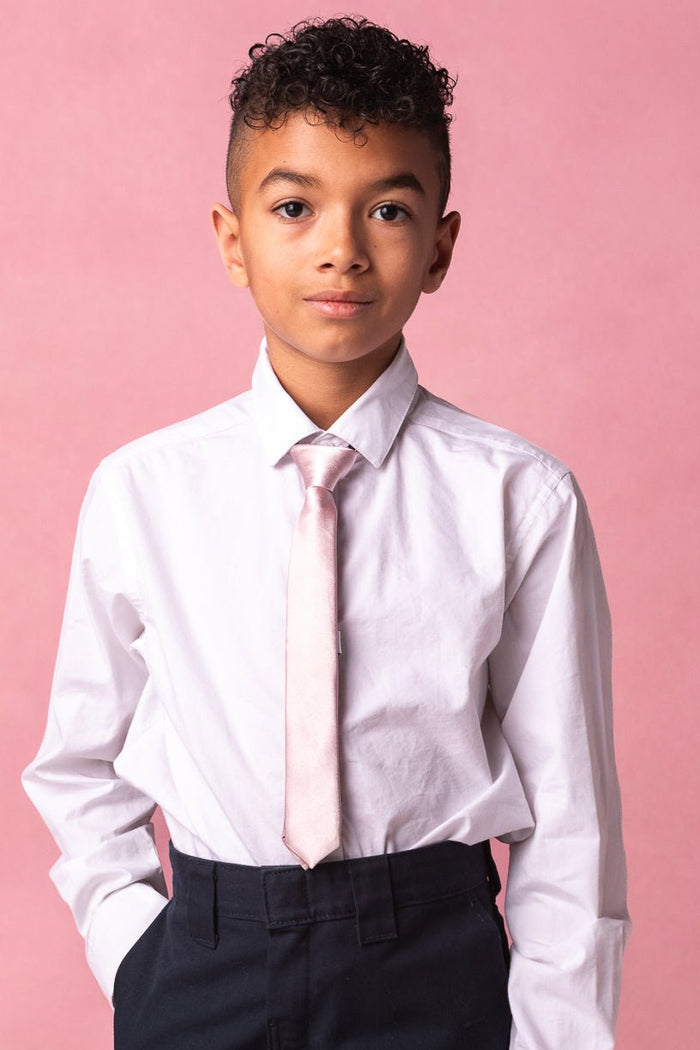 A young boy with short curly hair, dressed in a white dress shirt, dark pants, and the Boys Max Tie in Blush by Ivy City Co—a classic accessory that’s perfect for weddings—stands against a pink background with one hand in his pocket.