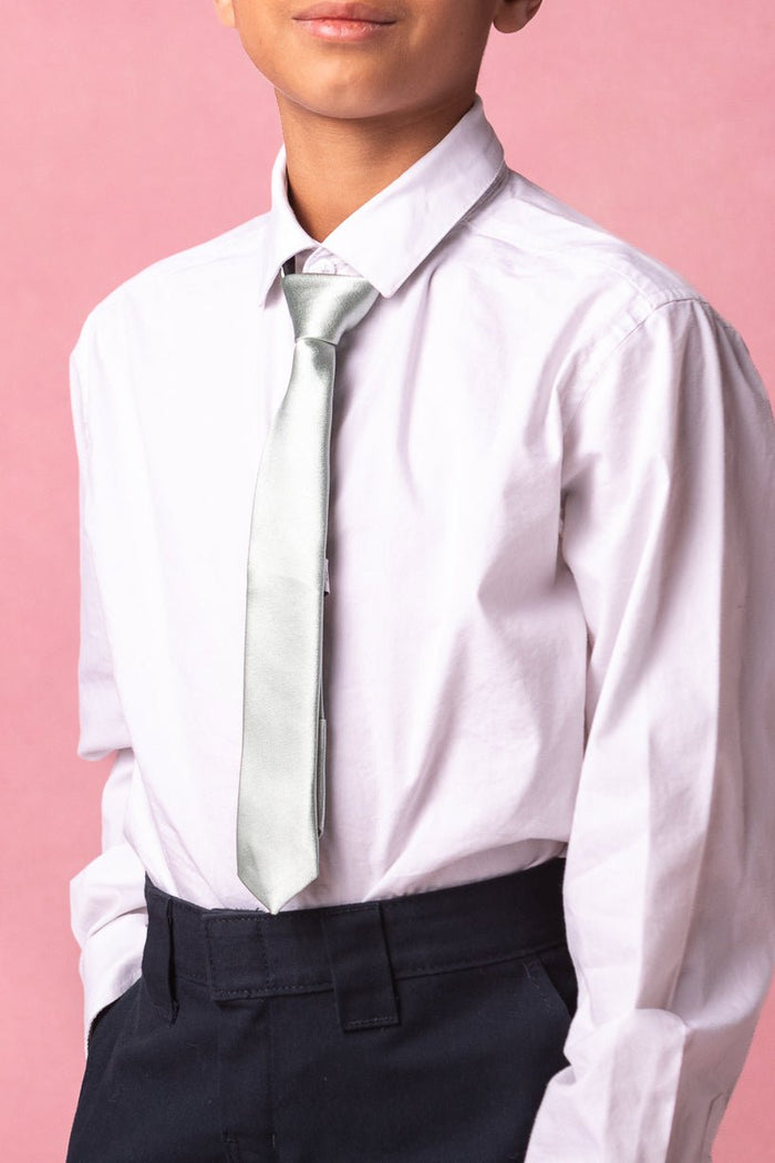 A person wearing a white dress shirt, dark pants, and the Ivy City Co Boys Max Tie in Sage stands against a pink background. The image is cropped at the shoulders and mid-thigh, keeping their full face out of view and drawing attention to the tie as an elegant wedding accessory.